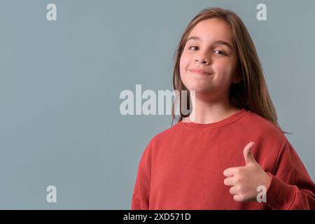 Ragazza di 11-12 anni che fa i pollici felici su. Indossa un maglione rosso che guarda la fotocamera con successo. Copia spazio Foto Stock