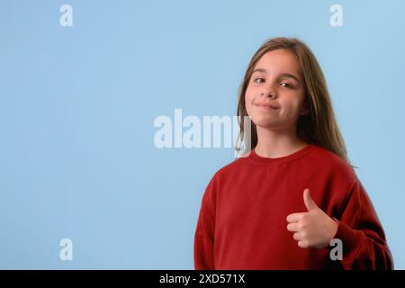 Una ragazza che fa felici i pollici. Indossa un maglione rosso che guarda la fotocamera con successo. Copia spazio Foto Stock