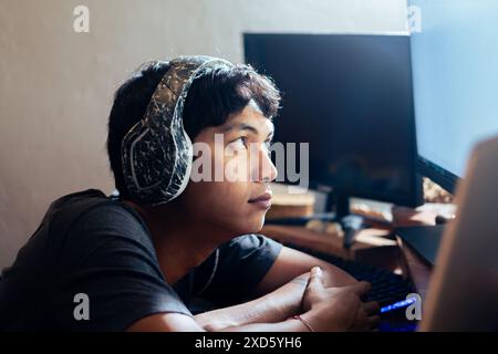 Un giovane uomo che indossa le cuffie guarda con attenzione lo schermo di un computer. È seduto a una scrivania con più monitor in una stanza poco illuminata. Foto Stock