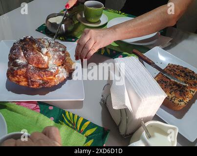 Spuntino con yogurt e pane al sesamo, ciambelle, tazza verde, tovaglioli, latte, cucchiaio, tovaglioli, zucchero e le mani di una donna che rompono la ciambella. Foto Stock