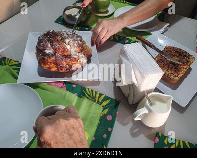 Spuntino con yogurt e pane al sesamo, ciambelle, tazza verde, tovaglioli, latte, cucchiaio, tovaglioli, zucchero e le mani di una donna che rompono la ciambella. Foto Stock