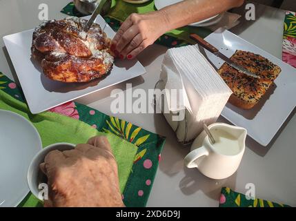 Spuntino con yogurt e pane al sesamo, ciambelle, tazza verde, tovaglioli, latte, cucchiaio, tovaglioli, zucchero e le mani di una donna che rompono la ciambella. Foto Stock