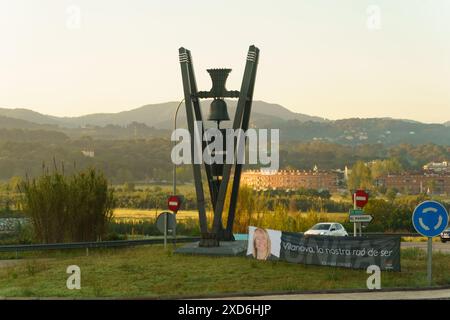 El Masnou, Spagna - 15 maggio 2023: Una scultura moderna con una grande campana si trova al centro di una rotonda a Villanova, in Spagna, con colline e costruzioni Foto Stock