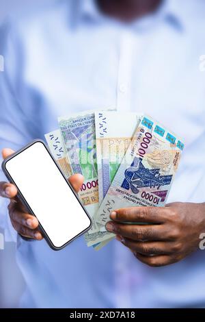 Persona nera che tiene in mano le note del franco CFA dell'Africa occidentale e mostra il telefono con schermo vuoto alla fotocamera. Mano che tiene banconote in valuta dell'Africa occidentale Foto Stock