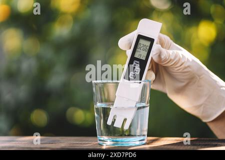 Tester per misuratore della qualità dell'acqua immerso in vetro per abbeveraggio, su sfondo naturale sfocato. Misurazione di impurità nocive, acqua distillata, acqua purificante Foto Stock