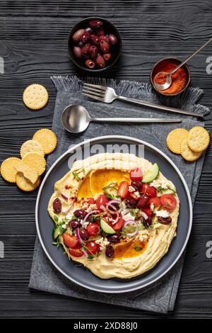 Hummus greco carico di cetrioli freschi, olive, pomodori, feta, cipolle rosse e timo su un tavolo di legno nero con cracker, cucchiaio e. Foto Stock