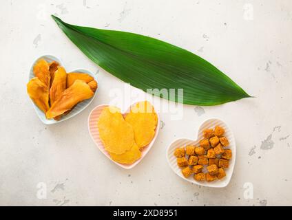 Fette di mango essiccate e palle in piatti a forma di cuore su sfondo chiaro con foglia Foto Stock