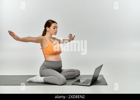 Una giovane donna incinta seduta su un tappetino yoga, che utilizza un computer portatile per il lavoro e il relax. Foto Stock