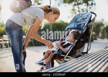 Una giovane madre offre una bottiglia d'acqua al suo bambino, che siede pazientemente in un passeggino, in una giornata di sole nel parco. Foto Stock