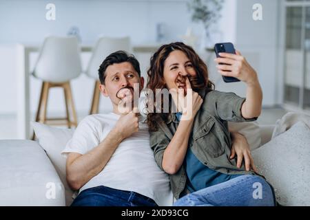 Coppia che crea volti divertenti con i baffi dei capelli, scatta divertenti selfie insieme a casa, cattura momenti di gioia e stupidità. L'uomo fa baffi divertenti dai capelli della donna, scatta foto sullo smartphone. Foto Stock