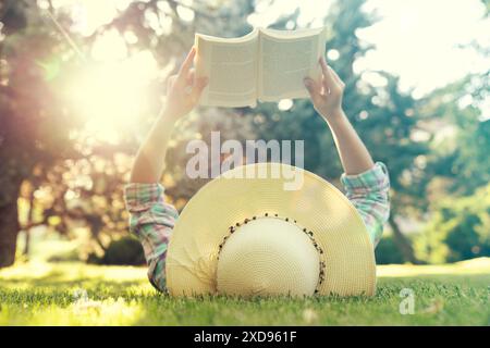 Una persona è sdraiata sull'erba con un libro in grembo. Una giovane donna con un cappello di paglia ama passare il tempo a leggere un libro nel parco Foto Stock