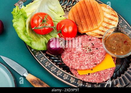 Hamburger freschi, formaggio, verdure e panini per la preparazione. Cotolette di bistecca crude con carne macinata. Foto Stock