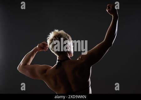 Ripresa retrovisiva di una donna sportiva vivace e forte in posa da competizione che solleva il braccio. Ripresa con retroilluminazione di una donna irriconoscibile bodybuilder in uno studio scuro con una luce spettacolare su sfondo nero, spazio di copia Foto Stock