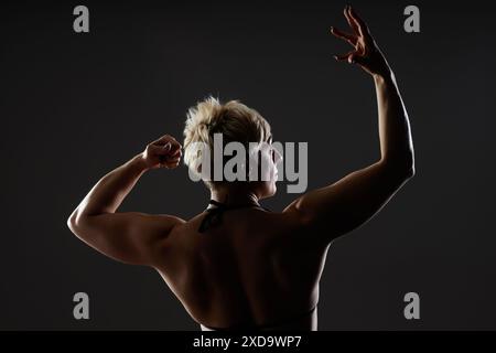 Ripresa con vista posteriore media di un'atleta libera che posa con le braccia in alto in modo artistico. Ritratto in controluce del bodybuilder che mostra i muscoli della schiena in uno studio scuro su sfondo nero, spazio di copia Foto Stock