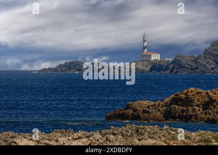 Splendida vista del faro di Faro de Favaritx arroccato sull'aspra costa rocciosa di Minorca, con un cielo spettacolare e un mare blu profondo. Foto Stock