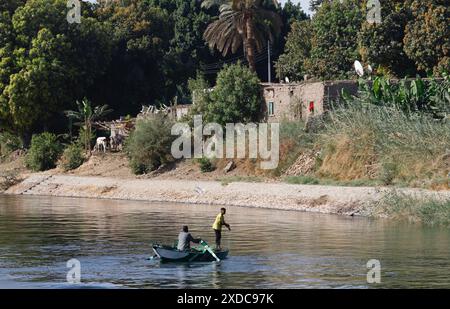 Un uomo e un ragazzo che pescano da una barca a remi nel Nilo di fronte a una casa in mattoni di fango con piatti satellitari e bestiame sulla fertile riva del fiume. Foto Stock