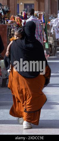 Nel souk di Assuan una donna egiziana in un hijab nero e un abaya arancione porta una bambina con un taglio di capelli pulito, anche lei con l'arancio, che dorme velocemente. Foto Stock