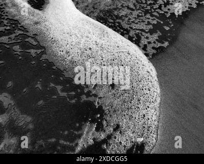 Una fotografia in bianco e nero che raffigura le onde schiumose dell'oceano su una spiaggia sabbiosa. L'immagine acquisisce un pattern astratto dinamico creato b Foto Stock