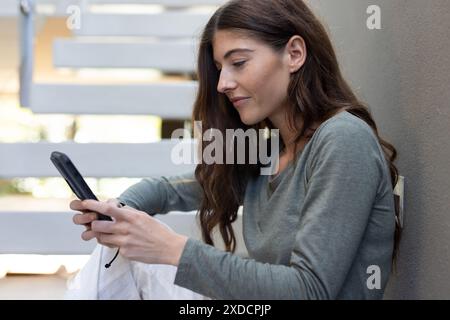 donna seduta sulle scale di casa, che utilizza lo smartphone e si concentra sullo schermo Foto Stock