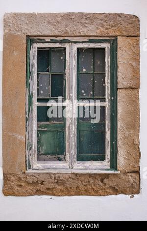 Vecchia finestra d'epoca di un antico edificio in pietra in Portogallo. Il telaio è sbiadito, il vetro manca nei punti e quelli che rimangono sono macchiati con pa Foto Stock