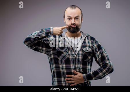 Uomo barbuto che tocca giocosamente la guancia, fa il viso divertente e le guance gonfiabili, emana umorismo e divertimento in un ambiente grigio da studio Foto Stock