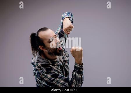 L'uomo furioso in camicia a quadri lancia pugni aggressivi su uno sfondo grigio, trasudando rabbia e dominio Foto Stock