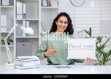 Ritratto di una giovane insegnante di inglese ispanica seduta in ufficio alla scrivania, tenendo un cartello tra le mani, puntando e sorridendo alla macchina fotografica. Foto Stock