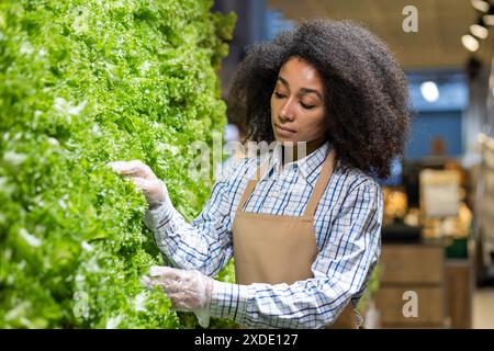 La giovane donna afroamericana in un grembiule uniforme e guanti lavora in un supermercato. si trova nel dipartimento di frutta e verdura fresca. Foto Stock