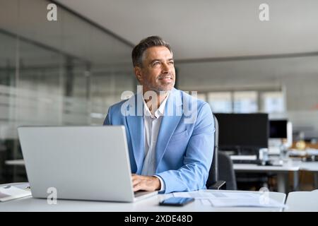 Un imprenditore felice di mezza età che lavora su un notebook che guarda lontano in ufficio. Foto Stock