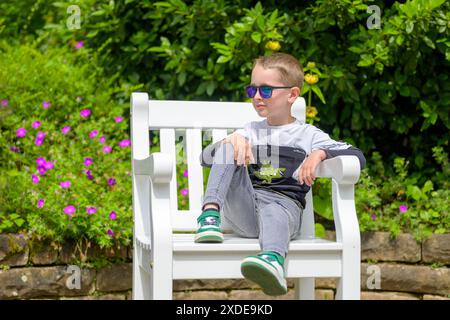 Un ragazzo che indossa occhiali da sole e abiti casual si rilassa su una panca bianca in un giardino vivace. Foto Stock