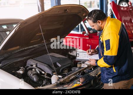 meccanico uomo che controlla il motore aperto cofano cerca elenco di controllo servizio di manutenzione auto in officina Foto Stock