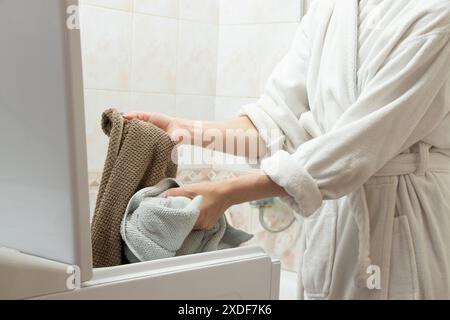 Una ragazza con l'abito bianco prende un asciugamano dalla lavatrice del bagno, lavando le cose, facendo i compiti Foto Stock