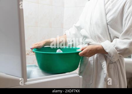 Una ragazza con l'abito bianco toglie le cose dalla lavatrice in bagno e le mette in una ciotola, lava le cose e fa i compiti Foto Stock