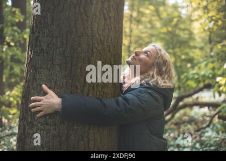 Una donna bionda con un lungo cappotto nero sorride e abbraccia un enorme albero nella natura selvaggia, simboleggiando una profonda connessione e l'impegno a proteggere il nostro mos Foto Stock