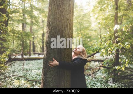 Una donna bionda con un lungo cappotto nero sorride e abbraccia un enorme albero nella natura selvaggia, simboleggiando una profonda connessione e l'impegno a proteggere il nostro mos Foto Stock