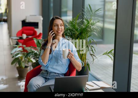 Una donna d'affari professionista è al telefono e si gusta un caffè in un ufficio moderno con un computer portatile Foto Stock