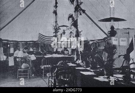 Questa fotografia in bianco e nero, scattata nel 1900, raffigura la sala lettura dell'ospedale da campo di Wynberg, in Sudafrica, durante la guerra boera. L'immagine cattura un momento di pace mentre gli individui si siedono ai tavoli, leggono o conversano, tra il caos e le difficoltà del tempo di guerra. L'ambientazione è una tenda con una grande bandiera americana in evidenza, suggerendo la presenza di forze americane nella regione. Foto Stock