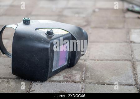 Primo piano di una maschera a casco per saldatura distesa su una superficie pavimentata con una persona che lavora su uno sfondo sfocato. Foto Stock