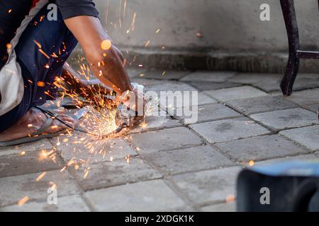 Un lavoratore che utilizza una smerigliatrice angolare per tagliare un oggetto metallico, causando scintille. Il lavoratore si sta inginocchiando su un pavimento piastrellato e indossa un panno casual Foto Stock