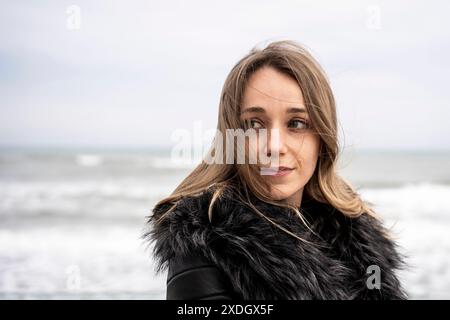 Donna con un cappotto di pelliccia che guarda con cura la spiaggia in una ventosa giornata invernale. Guarda in lontananza con il mare e le onde sullo sfondo, si conva Foto Stock