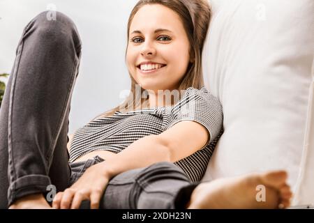 Una giovane donna siede comodamente su un divano bianco a casa, rilassandosi con un sorriso, incarnando il benessere in un sincero momento di gioia Foto Stock