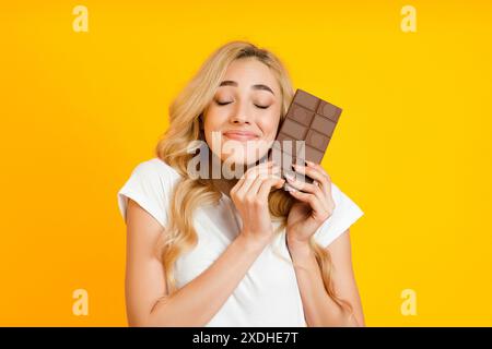 Donna felice bionda che si gusta Chocolate Bar su sfondo giallo, primo piano Foto Stock