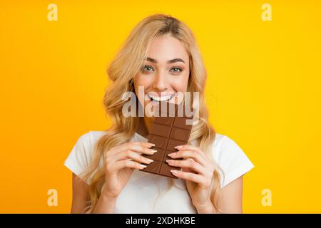 Happy Blonde Woman che mangia Chocolate Bar su sfondo giallo Foto Stock