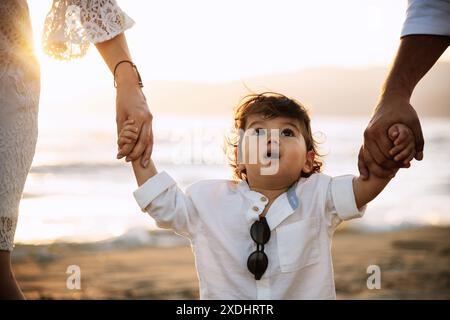 Un bambino di un anno con una camicia bianca e occhiali da sole cammina sulla spiaggia, tenendo le mani dei suoi genitori. Lo sfondo presenta sabbia e la calda luce di Foto Stock