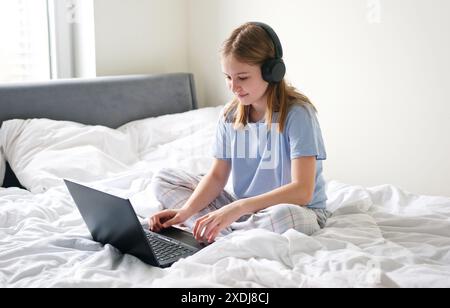 Dolce ragazza seduto a letto, indossando le cuffie e aprendo il laptop per fare i compiti online Foto Stock