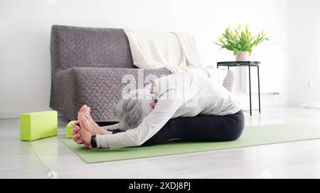 Donna attiva anziana che fa yoga piega posa a casa Foto Stock