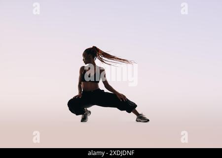 La donna della generazione Z con i capelli intrecciati mostra il suo atletismo in uno studio. Dimostra flessibilità mentre posa, si allunga e accovaccia con grazia. H Foto Stock