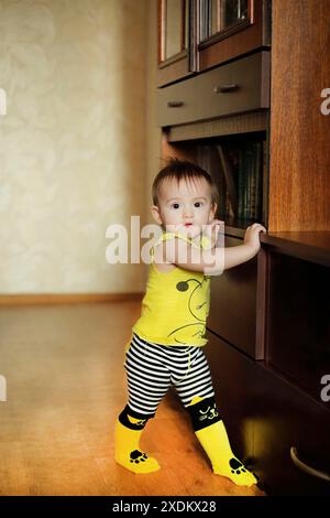 Un bambino si trova vicino a mobili di legno, indossando una camicia gialla e pantaloncini a righe in una stanza illuminata con un'atmosfera giocosa Foto Stock