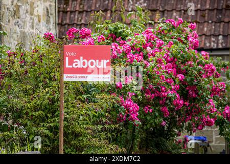 WARMINSTER, WILTSHIRE, Regno Unito, 23 giugno 2024, firma la pubblicità del partito politico laburista. Segno del Partito Laburista rosso che dice "Vota laburista", su un palo di legno di fronte a un cespuglio di graziose rose rosse. Pubblicità politica in vista delle elezioni generali. Accreditamento John Rose/Alamy Live News Foto Stock