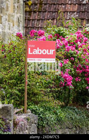 WARMINSTER, WILTSHIRE, Regno Unito, 23 giugno 2024, firma la pubblicità del partito politico laburista. Segno del Partito Laburista rosso che dice "Vota laburista", su un palo di legno di fronte a un cespuglio di graziose rose rosse. Pubblicità politica in vista delle elezioni generali. Accreditamento John Rose/Alamy Live News Foto Stock